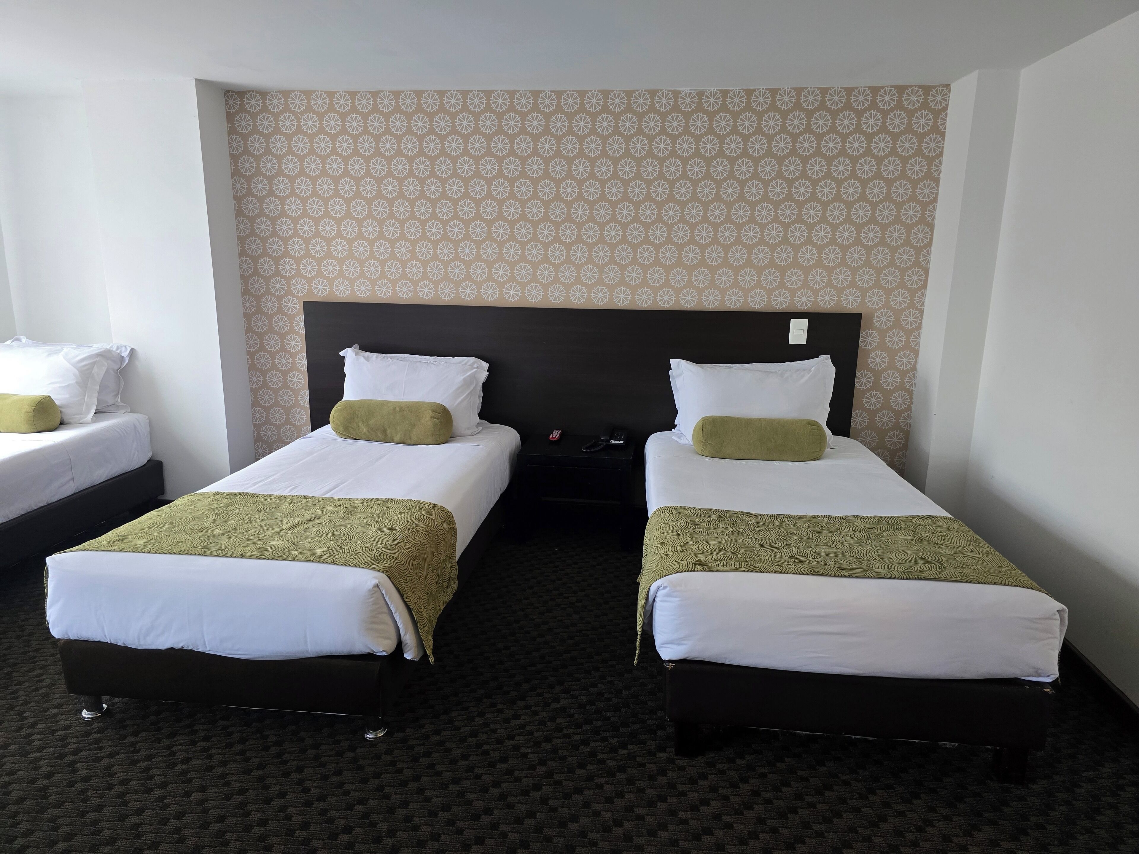 A hotel room with two single beds, each with white bedding and green throw pillows. The room has a patterned wallpaper and a dark headboard.