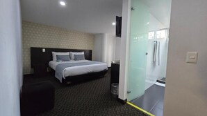 Une chambre d’hôtel moderne dotée d’un grand lit, d’un petit coin salon et d’une paroi en verre qui sépare la chambre de l’espace douche.