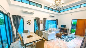 A spacious living area with a dining table, sofas, and a TV. There are large windows and an air conditioning unit on the wall.