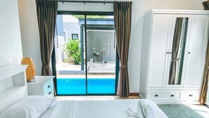 A bedroom with a bed, a nightstand, a lamp, a wardrobe, and a view of a swimming pool.