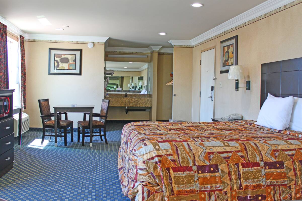 Une chambre d’hôtel avec un grand lit, un bureau avec deux chaises, un miroir et une salle de bain visible à travers une porte ouverte.