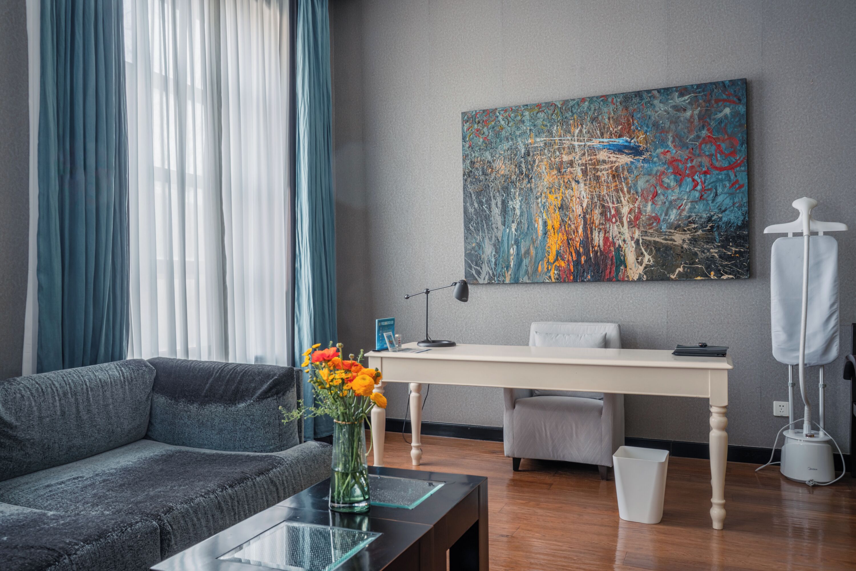 A modern living room with a grey sofa, a glass coffee table, a white desk, and a colorful abstract painting on the wall.