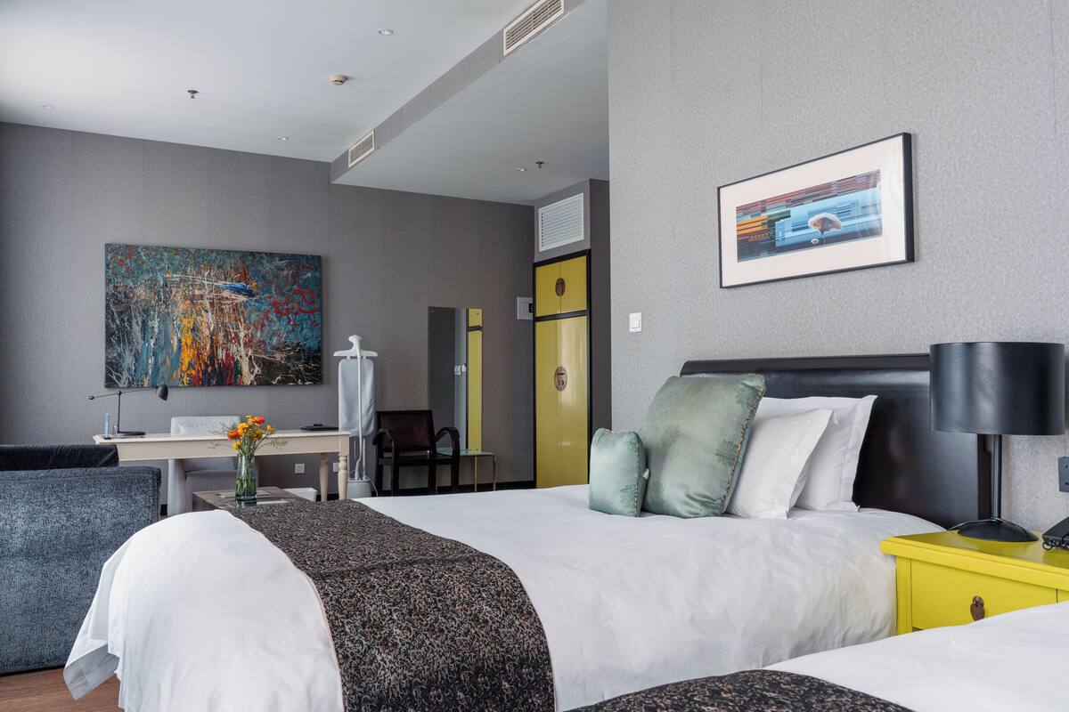 A modern hotel room with a large bed, a desk, and a chair. There is a painting on the wall and a window with blinds.