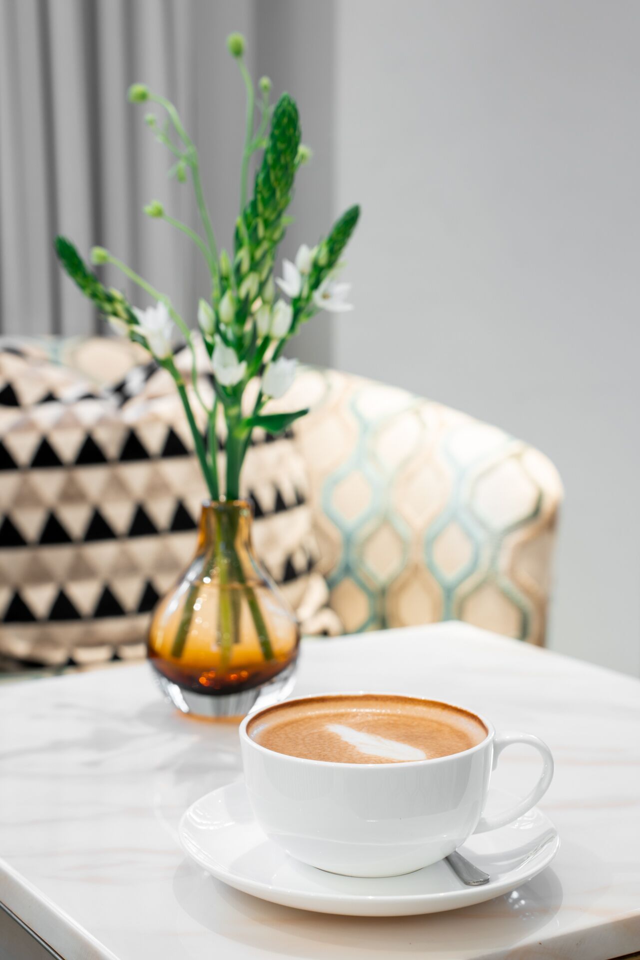 A white cup of coffee on a white saucer on a marble table. In the background, there is a patterned cushion and a vase with green plants.