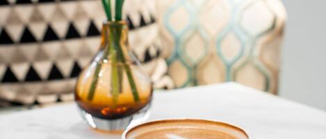 A white cup of coffee on a white saucer on a marble table. In the background, there is a patterned cushion and a vase with green plants.