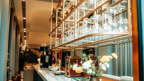 Un área de bar con estantes llenos de vasos y botellas, un jarrón con flores y vista al interior.