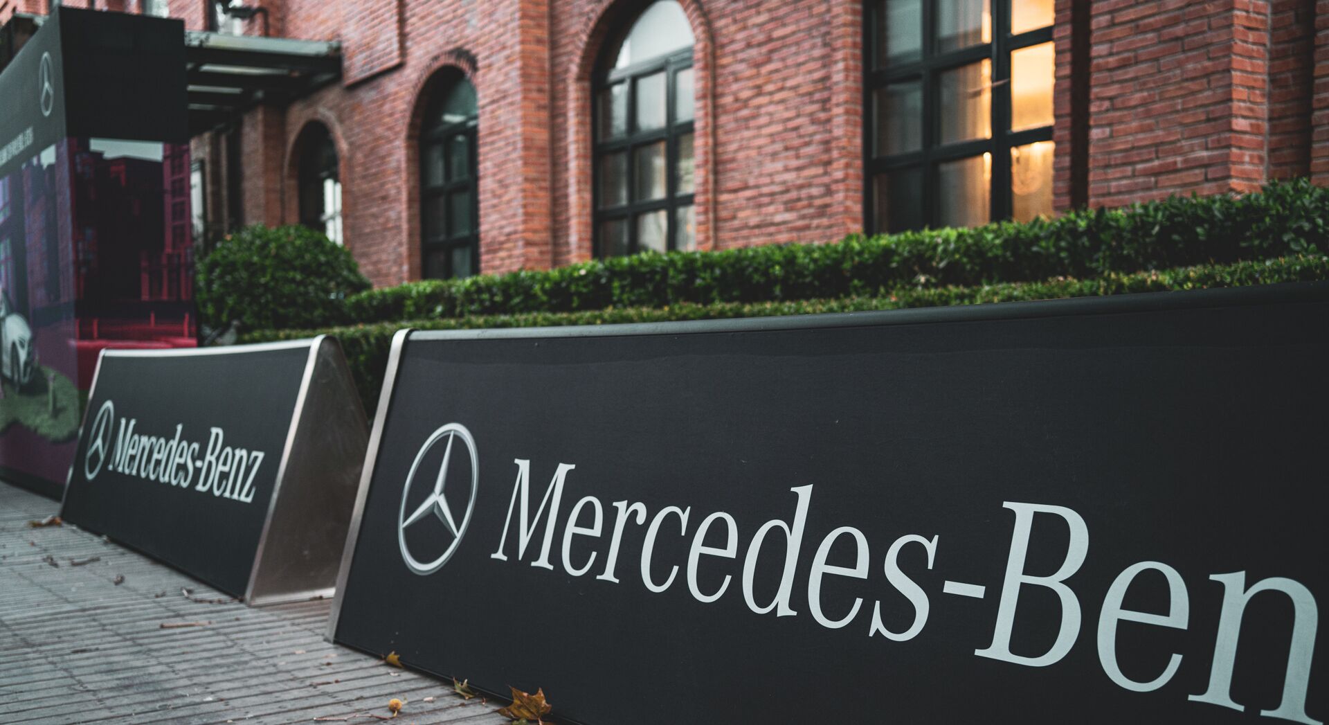 Un letrero negro con el logotipo y el nombre de Mercedes-Benz, ubicado al aire libre frente a un edificio de ladrillo con ventanas de arco.