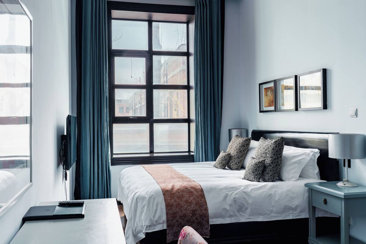 A hotel room with a large window, a bed with white bedding and a brown throw, a bedside table with a lamp, and a wall-mounted TV.