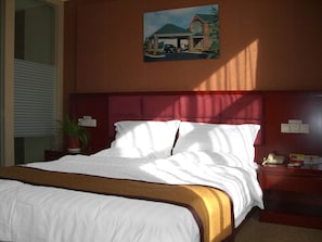 Habitación de hotel con una cama grande, cabecera de madera, un cuadro de un edificio en la pared, una mesita de noche con una lámpara y una planta en maceta.