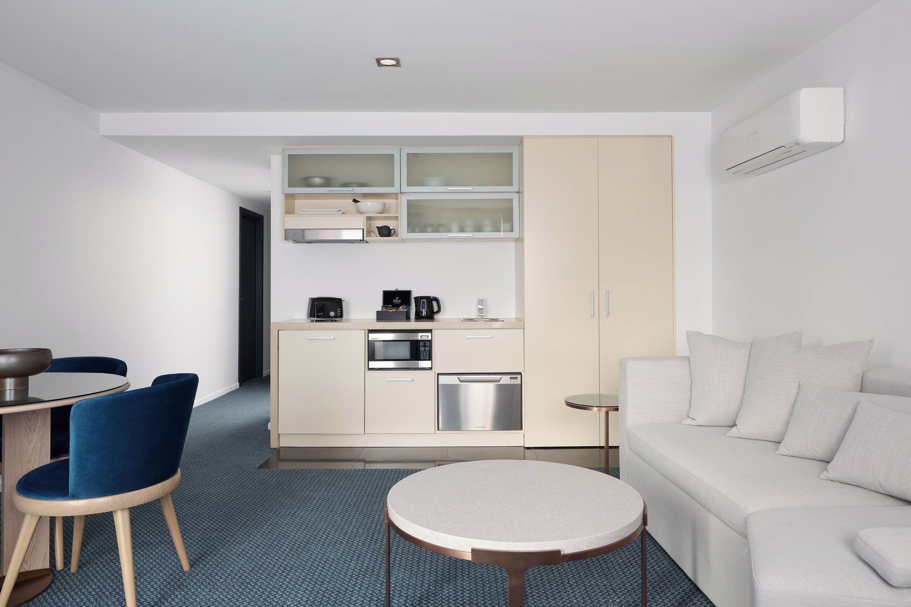Une chambre d’hôtel moderne et compacte, dotée d’une kitchenette, d’un coin repas avec des chaises bleues, d’une table ronde et d’un canapé.