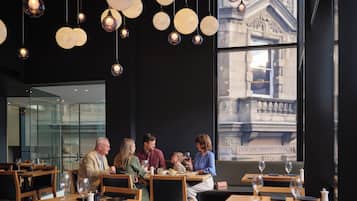 Een restaurant met ronde hanglampen, houten tafels en uitzicht op een gebouw door grote ramen.