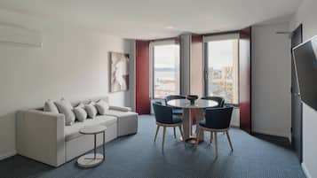 Una habitación de hotel moderna con un sofá gris, una mesita redonda y vistas a la ciudad a través de grandes ventanales.