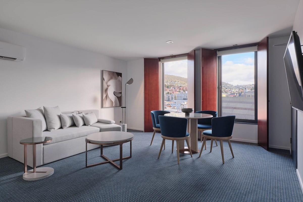 A modern hotel room with a sofa, two round tables, and chairs, a large window with a city view, and a staircase.