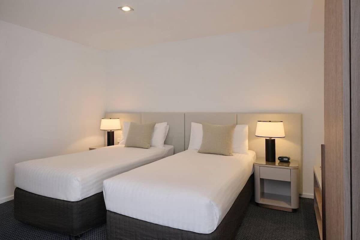 A hotel room with two single beds, bedside tables with lamps, and a small nightstand.
