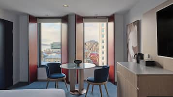 A hotel room with a bed, a round table with two chairs, a desk, and a large window offering a city view.