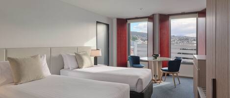 Une chambre d’hôtel avec deux lits, une petite table avec une chaise, une vue sur la ville et une fenêtre avec des rideaux rouges.