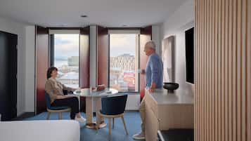 A hotel room with a bed, a round table with chairs, a desk with a bowl, and a large window offering a city view.