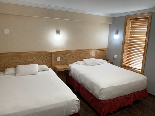 A hotel room with two beds, a wooden headboard, a small bedside table, a nightstand, a wall-mounted light, and a window with blinds.