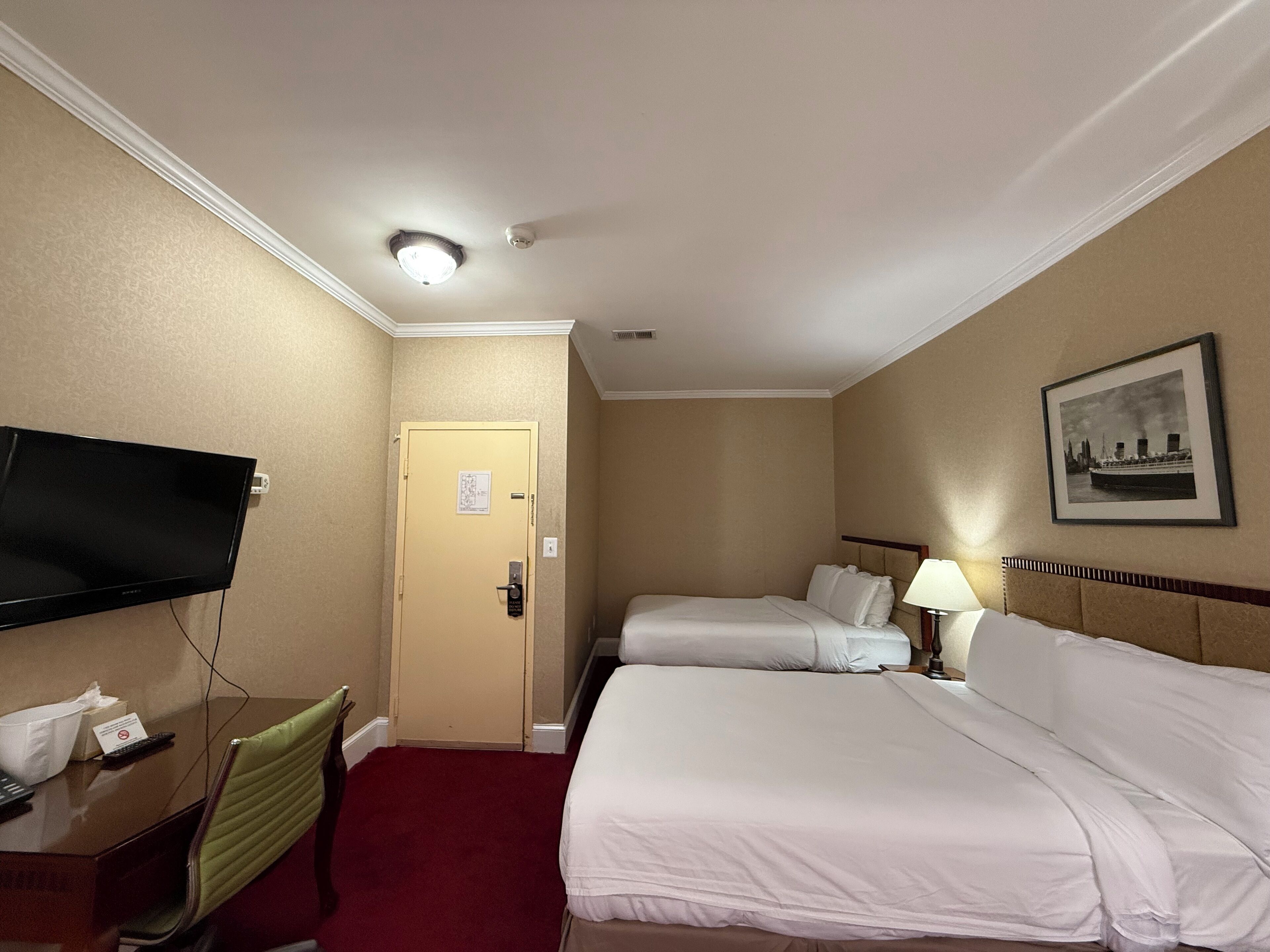 Een hotelkamer met twee bedden, een bureau met een televisie, een stoel, een deur en een lijst aan de muur.