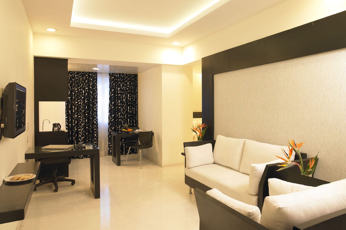 A modern hotel room with a sofa, a desk with a computer, a chair, and a dining area with a table and chairs.