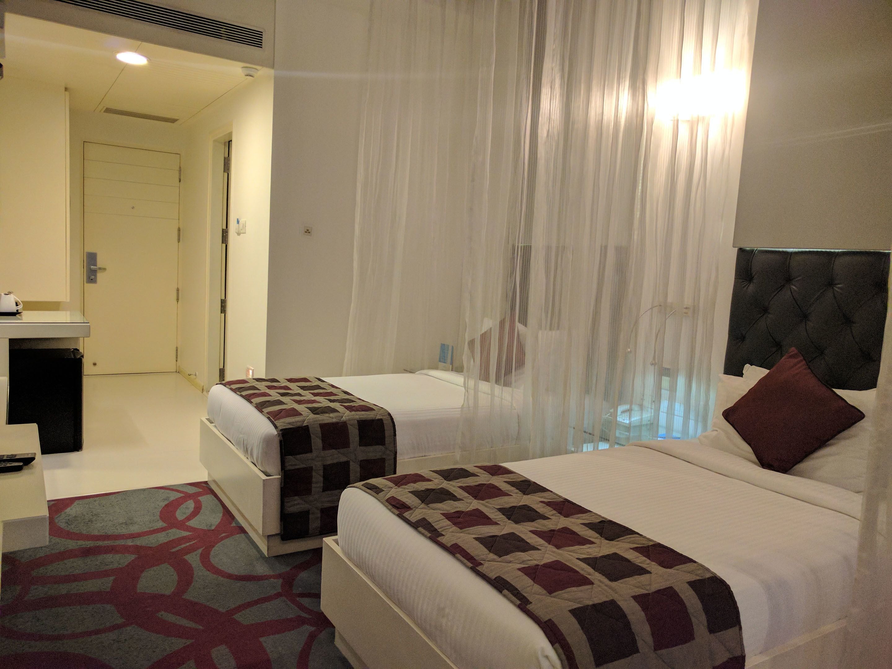 A hotel room with two beds, a bedside table, a mirror, and a window with sheer curtains.