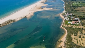 An aerial view of a coastal area with a beach, clear waters, and a residential area with houses and greenery. - Hotel Apolo (Vila Real Santo Antonio)