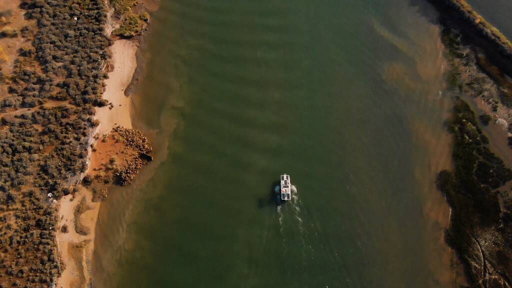Vista aérea de un barco sobre un agua tranquila, rodeado de vegetación escasa y una orilla arenosa.