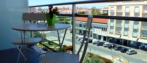 A balcony with a table and chairs, a potted plant, and a view of a street with parked cars and buildings.