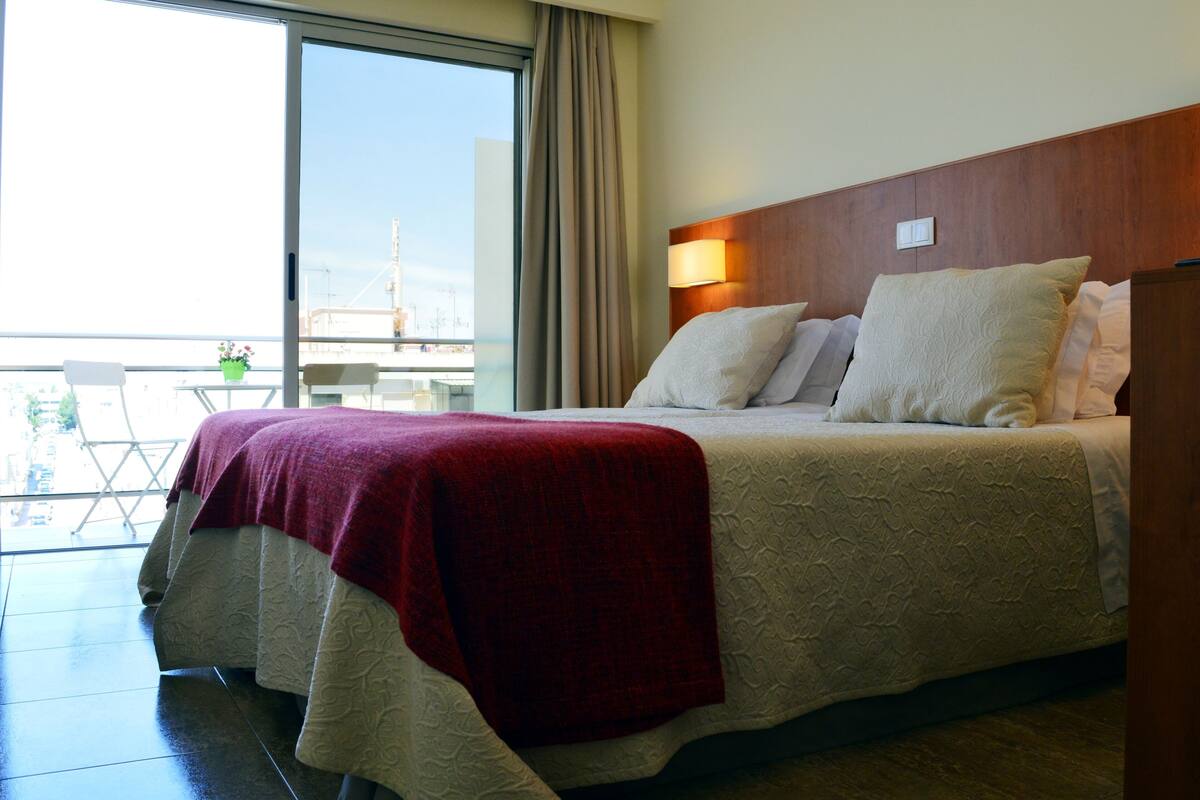 A hotel room with a large bed, a wooden headboard, and a view of the city through a sliding glass door.