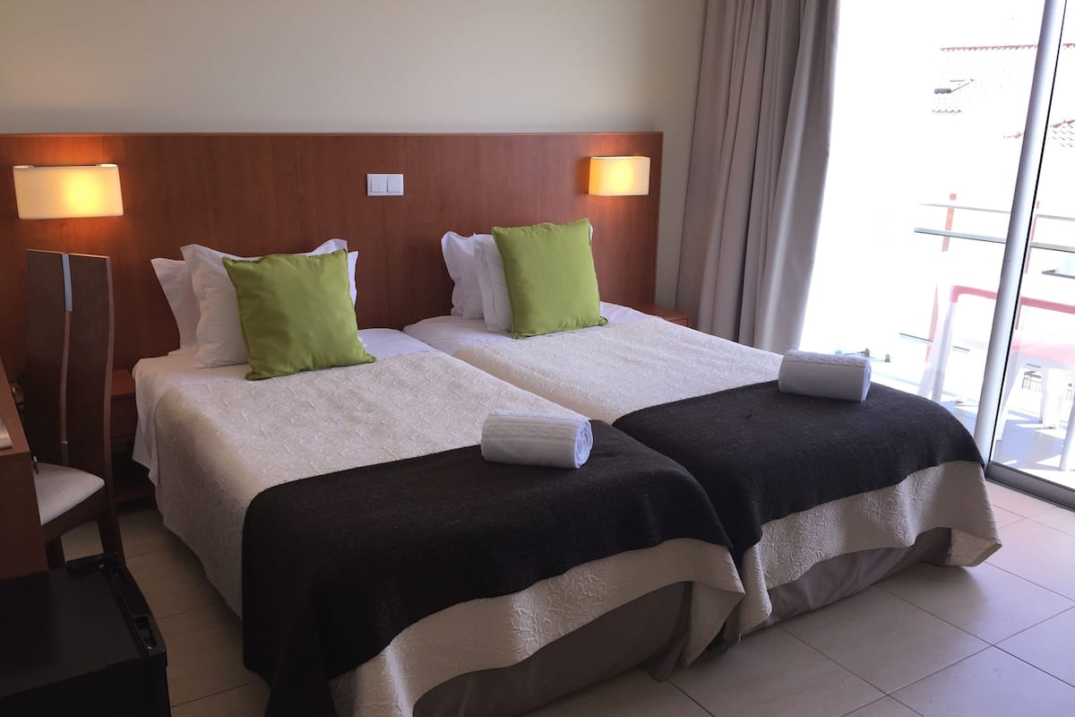 A hotel room with two beds, each with a white and green pillow, a wooden headboard, and a nightstand with a lamp.