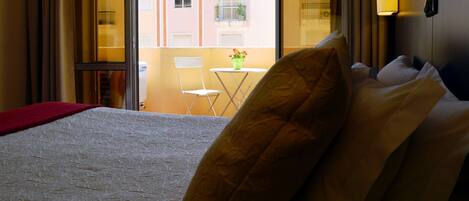 A hotel room with a bed, a chair, a small table with a vase, and a balcony with a view of buildings.