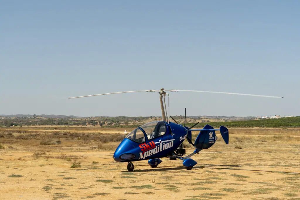 A blue helicopter with the text "SKU Expedition" on its side, parked on a dry, grassy field with a clear sky in the background.