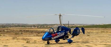 A blue helicopter with the text "SKU Expedition" on its side, parked on a dry, grassy field with a clear sky in the background.