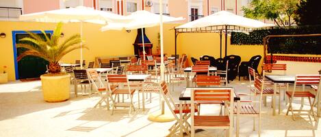 An outdoor seating area with tables and chairs under white umbrellas, surrounded by a yellow building with balconies.