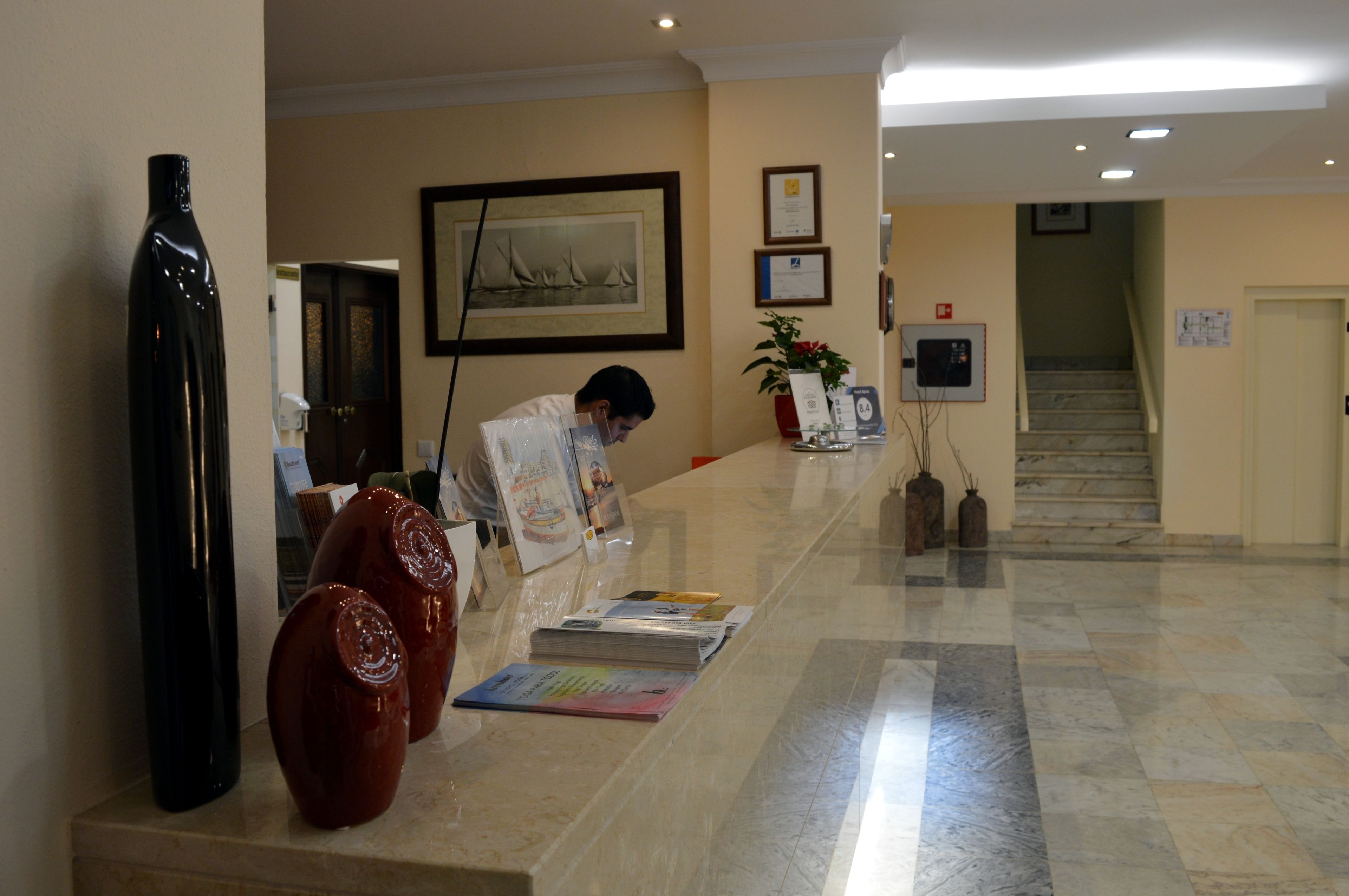 Zona de recepción de un hotel con suelo de mármol, una recepcionista, una obra de arte enmarcada y una escalera al fondo.