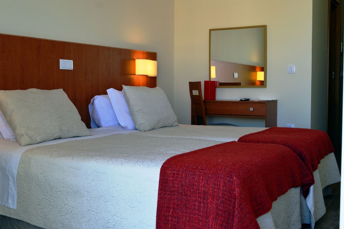 A hotel room with a large bed, a wooden headboard, a bedside table, a mirror, and a red throw blanket.
