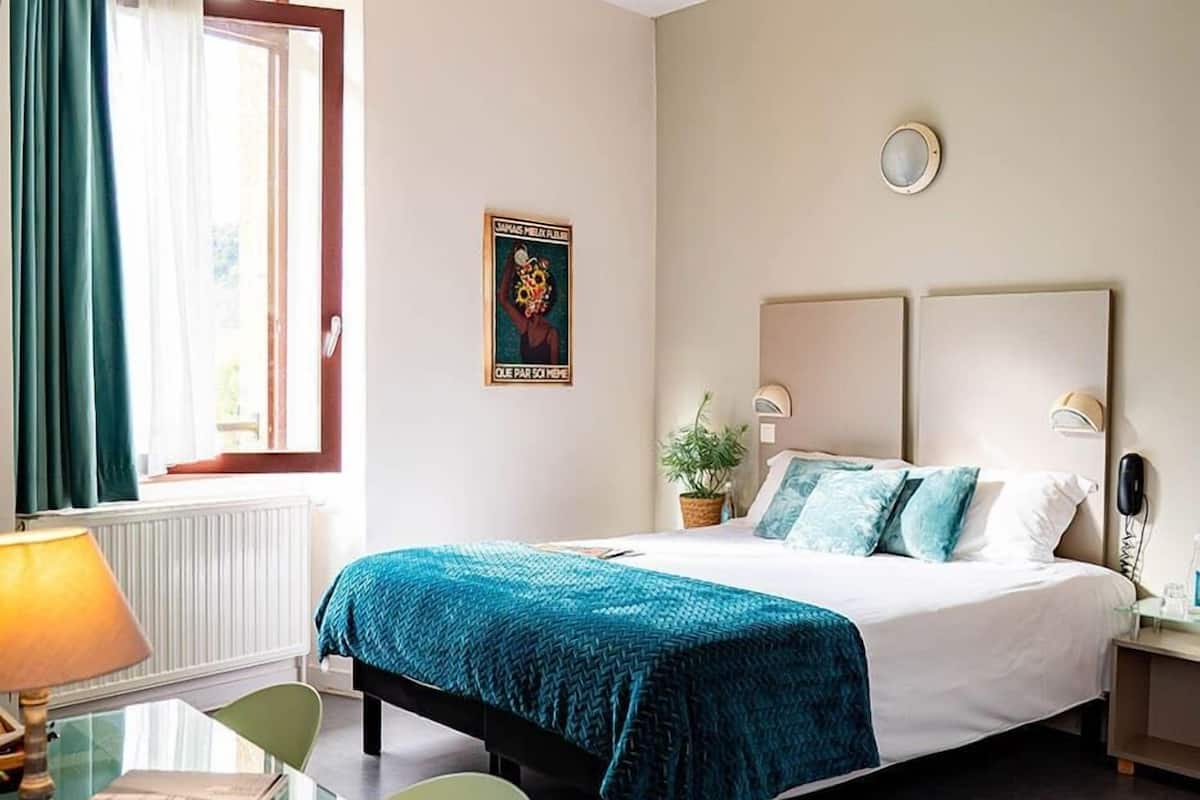 Habitación de hotel con cama, escritorio con lámpara, silla, ventana con cortinas y un reloj en la pared.