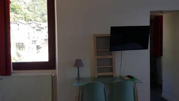 A small room with a window, a table, two chairs, a lamp, a TV on the wall, and a radiator.