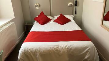 Une chambre d’hôtel comprenant un lit, deux tables de chevet, une petite table, un téléphone fixé au mur et une fenêtre avec des stores.