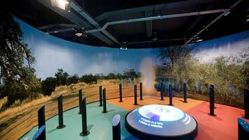 A circular interactive display with a sign in Spanish, surrounded by black poles, set against a backdrop of a painted landscape with trees and a fence.