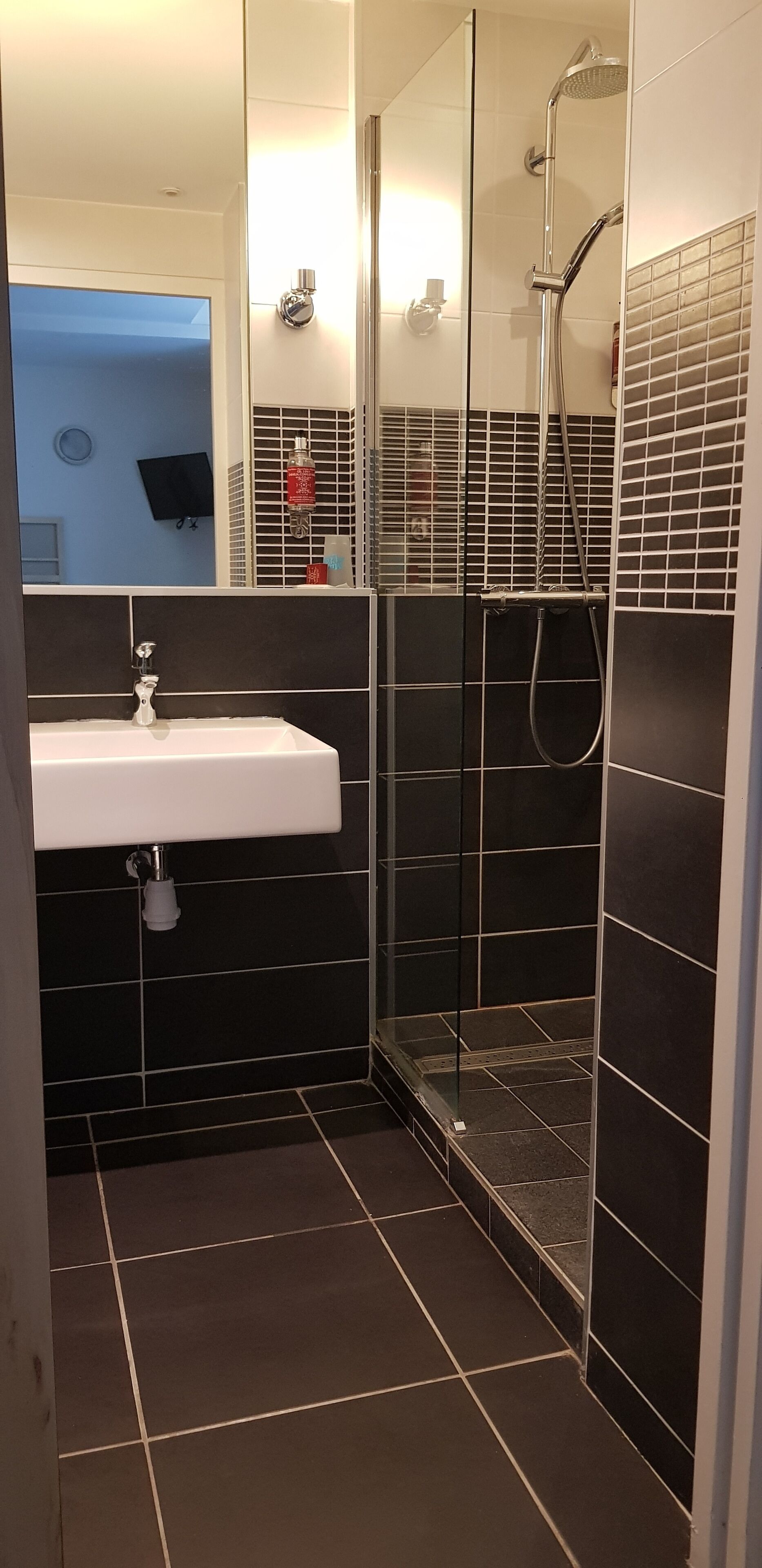 Ein modernes Badezimmer mit einem weißen Waschbecken, schwarzen Fliesenwänden und einer Duschkabine aus Glas.