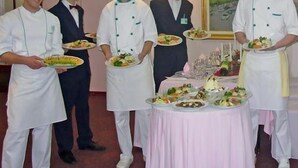Servicemedarbejdere i hvide uniformer og hatte bærer tallerkener med mad i et spiseområde med et bord dækket til et måltid.