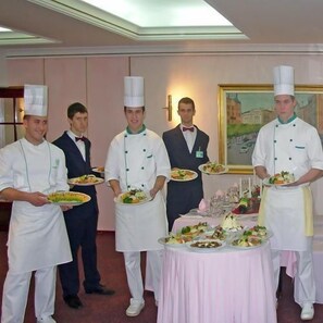 A group of waitstaff in white uniforms and hats holding plates of food in a dining area with a table set for a meal. - Palace Hotel (Belgrade)