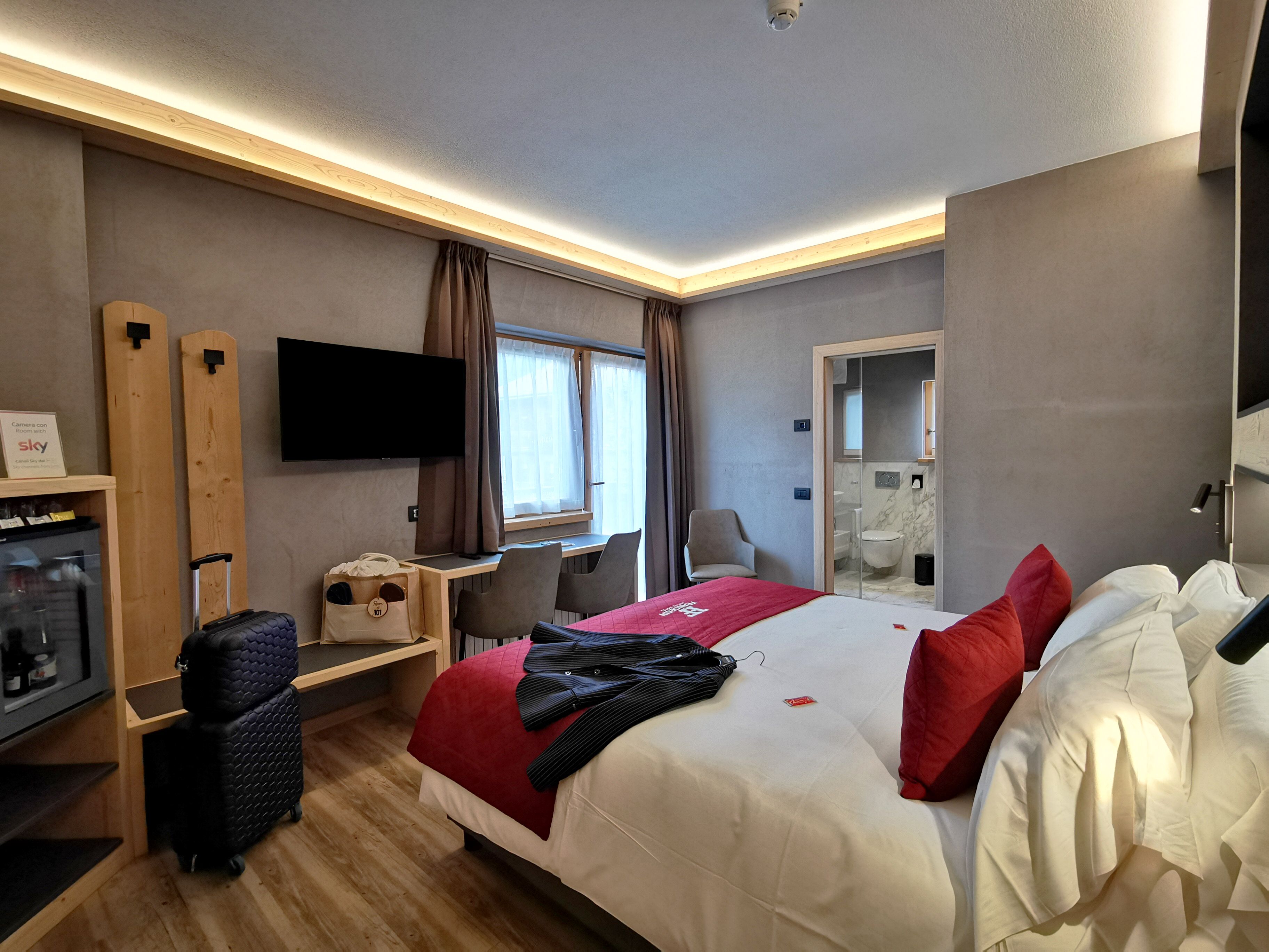 A hotel room with a bed, a desk, a chair, a television, a wardrobe, and a bathroom visible through an open door.
