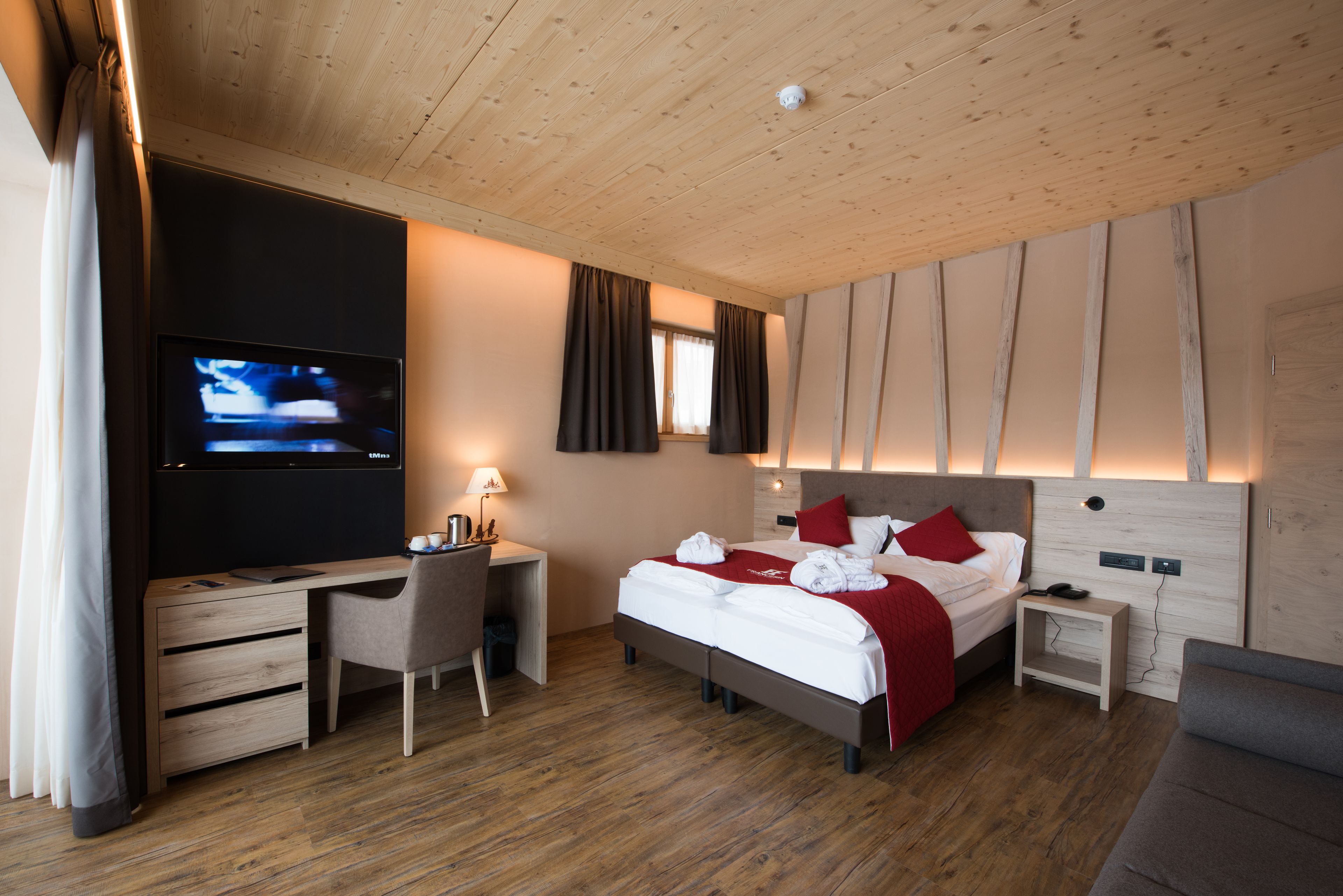 Una habitación de hotel moderna con techo de madera, un televisor de pantalla plana, un escritorio con silla, una cama con ropa de cama roja y una mesita de noche pequeña.