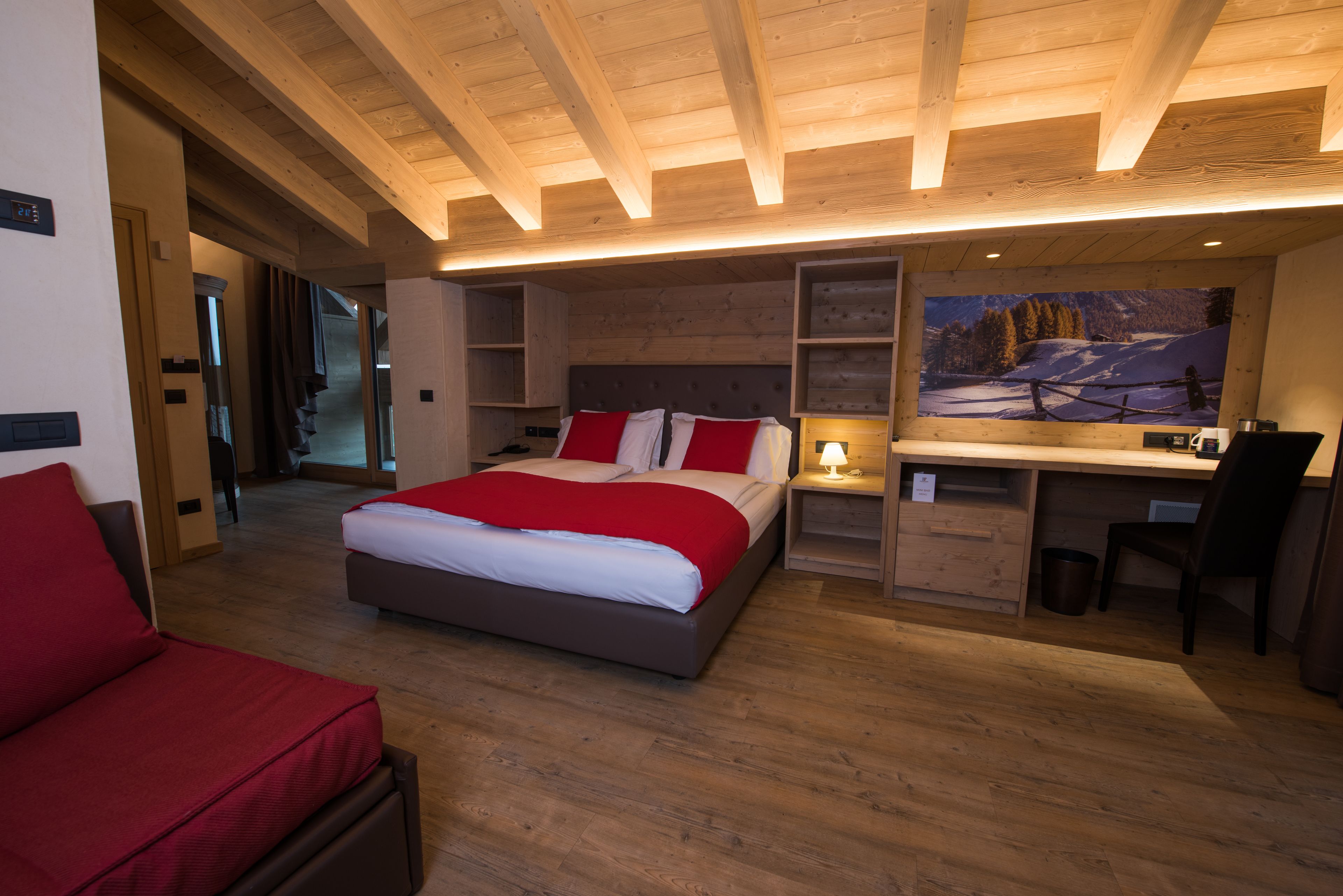 A modern bedroom with a large bed, a desk, and a chair. There is a painting on the wall and a wooden ceiling.