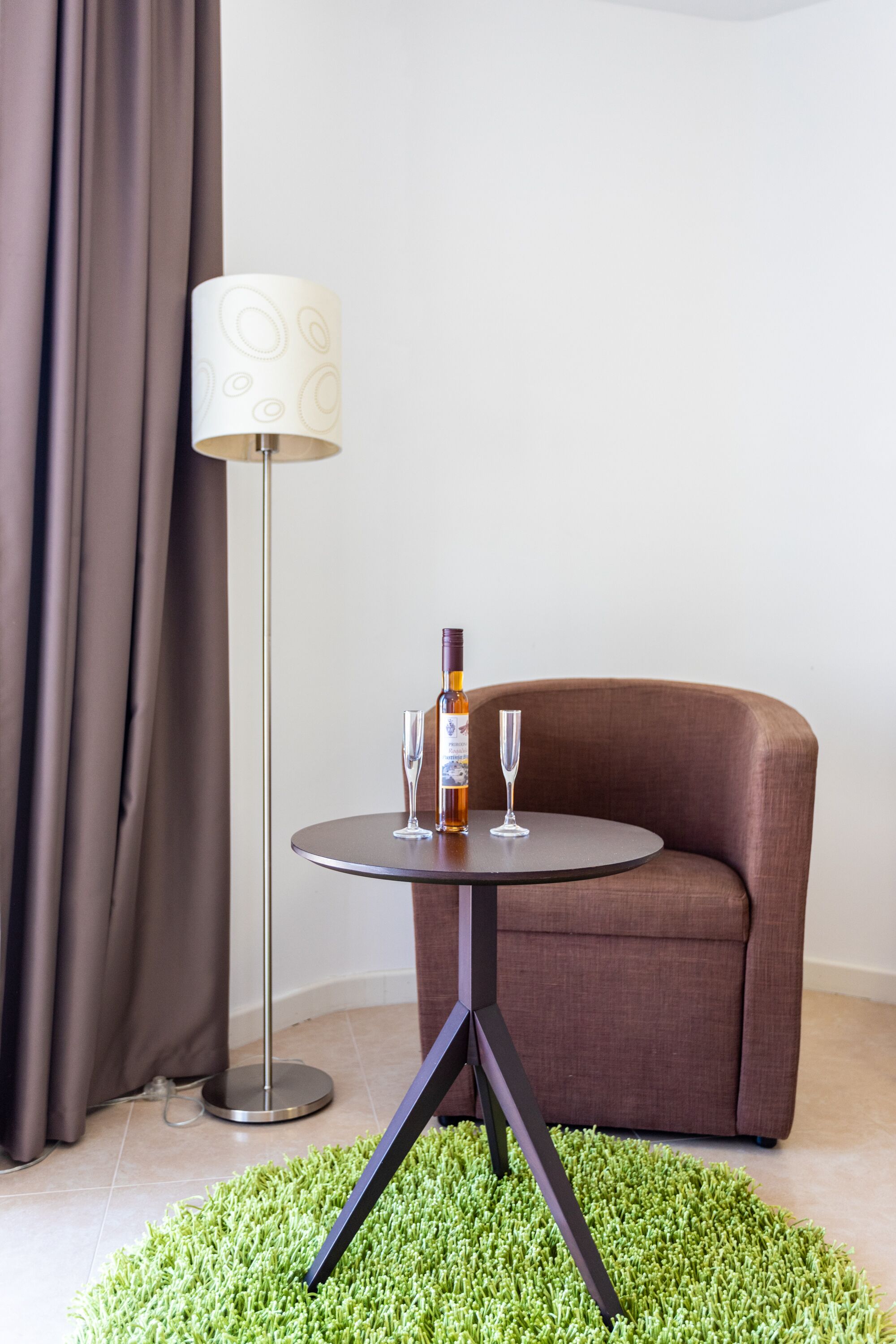 A round table with a bottle and two glasses, a brown sofa, a floor lamp, and a green carpet.