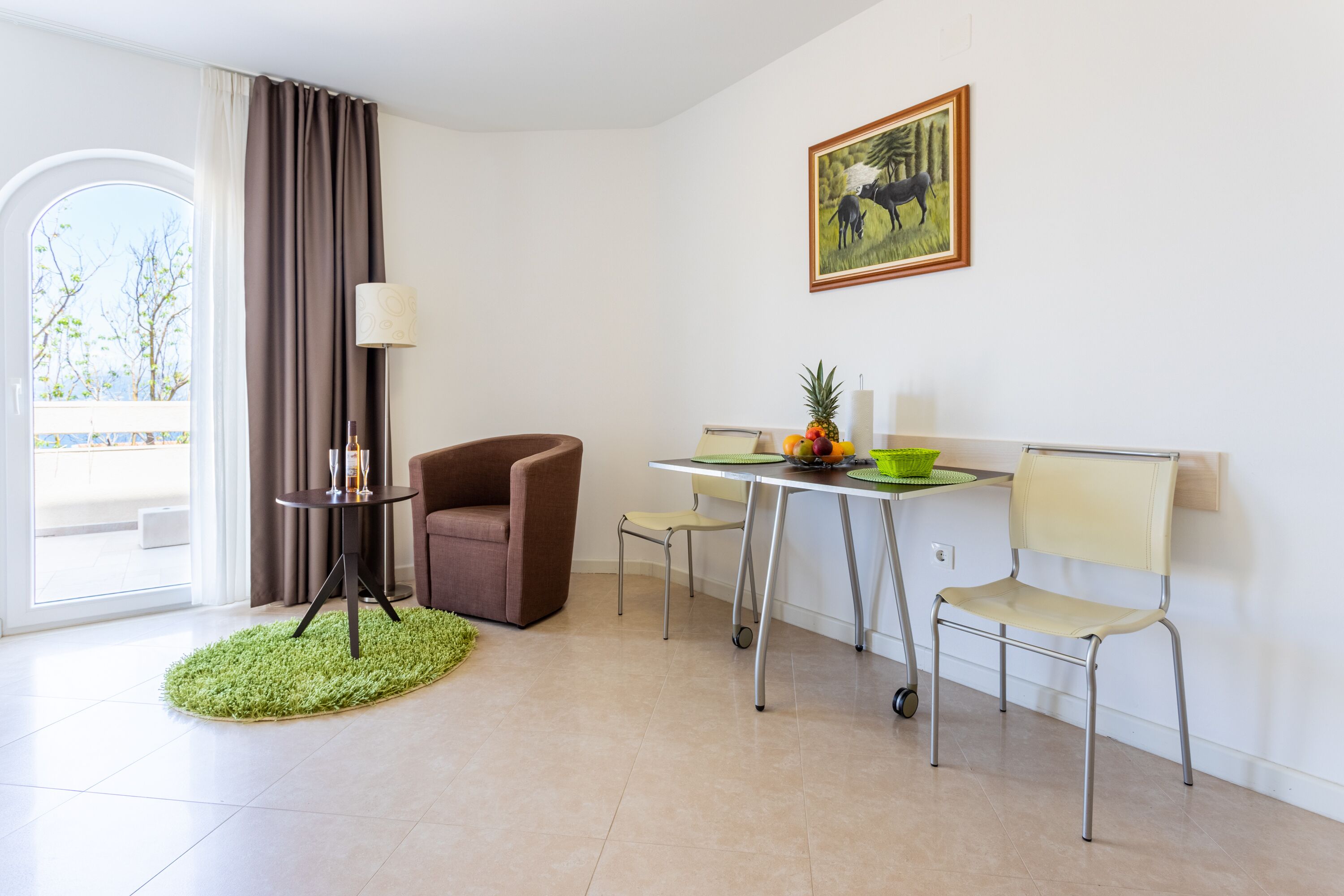 A modern room with a dining area, a brown armchair, a small table with a lamp, and a painting on the wall.