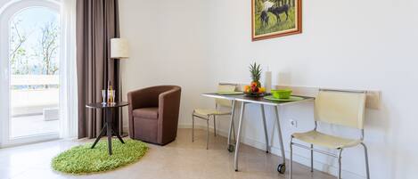 A modern room with a dining area, a brown armchair, a small table with a lamp, and a painting on the wall.