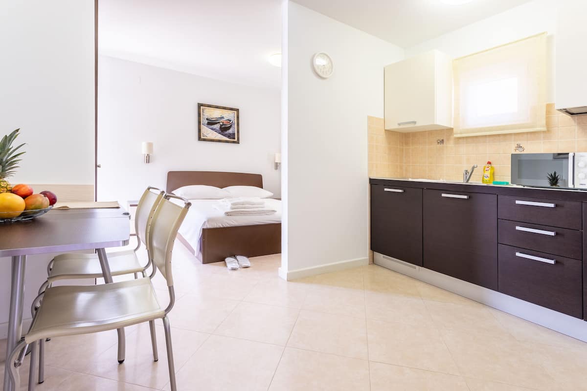 A hotel room with a bed, a dining area with a table and chairs, a kitchen with cabinets and a microwave, and a fruit bowl on the table.
